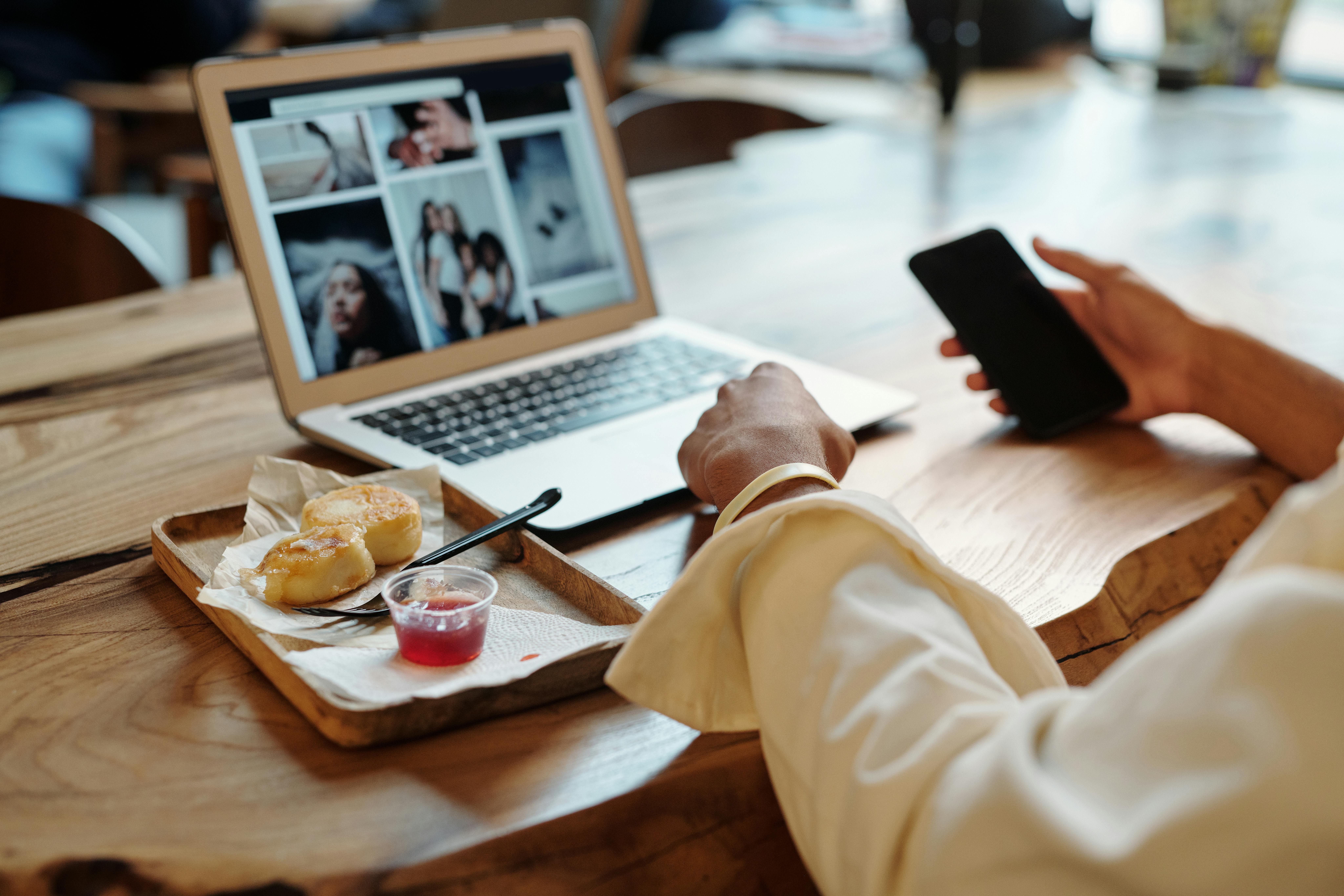 A person working remotely using a laptop and smartphone, with a snack on a wooden table.
